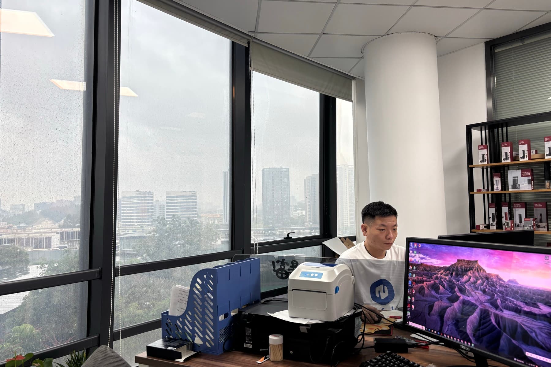 A Neofulfill team member working at a desk in the Shenzhen office, with a label printer and shelved products behind.